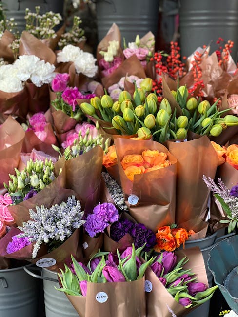 An assortment of fresh flowers, including pink and white roses, purple statice, and orange tulips, all wrapped in brown kraft paper. The bouquets are arranged in metal buckets, ready for flower delive