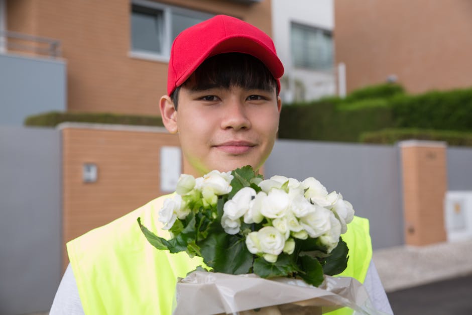 A young male flower delivery courier wearing a red cap and neon yellow vest holding a bouquet of fresh white roses wrapped in white paper, with green leaves visible, outdoors in front of modern reside