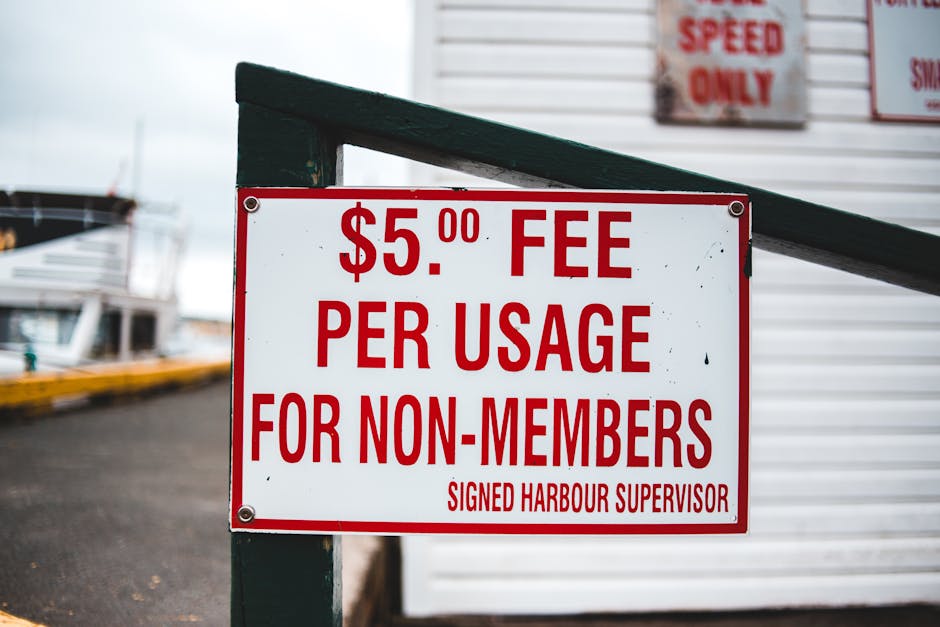 A white rectangular sign with bold red text indicating a $5.00 fee per usage for non-members, attached to a green metal railing at a harbor or marina. The background includes a blurred view of docked